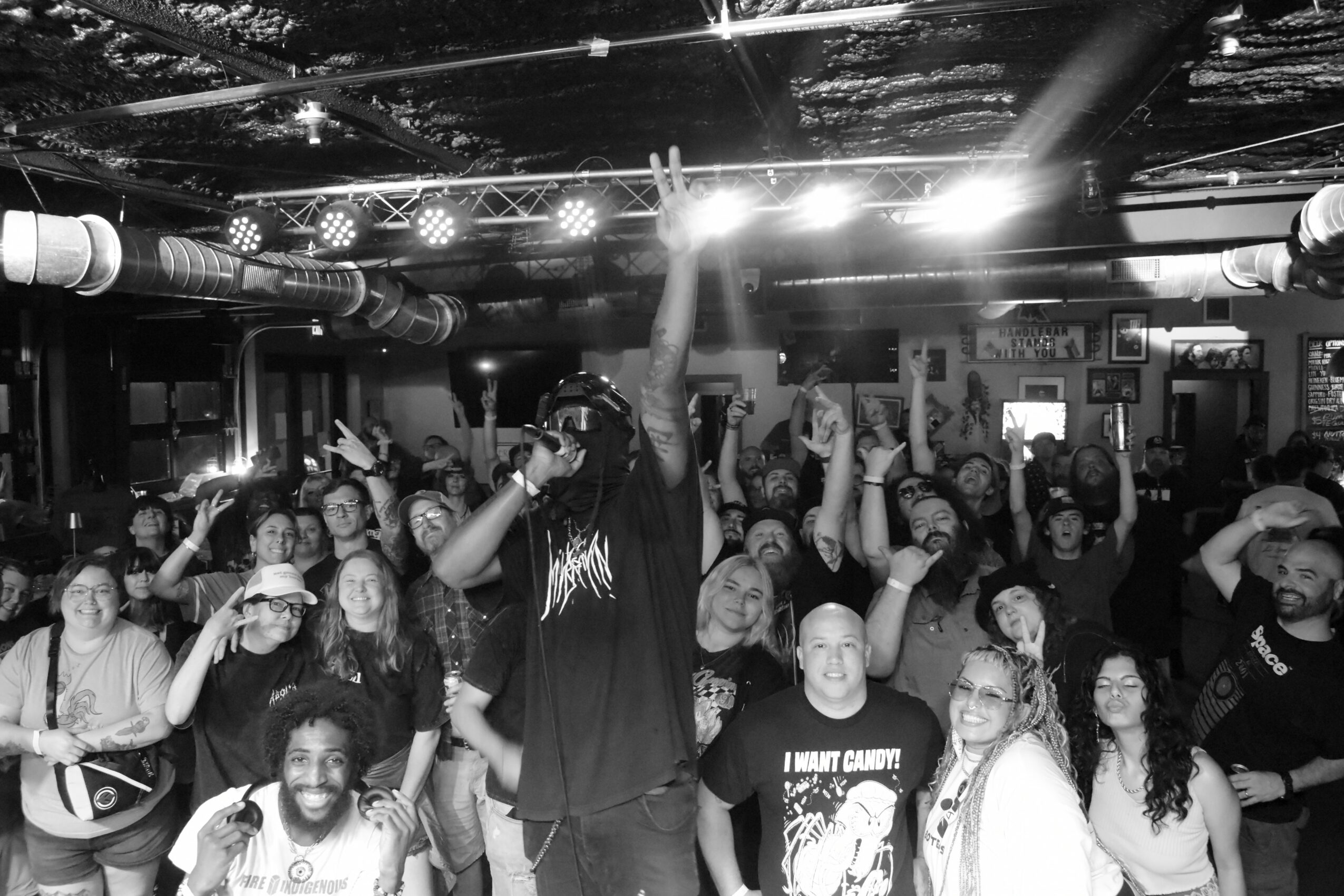 Crowd of people at a concert or event, some raising hands, with stage lighting overhead, in a lively indoor setting.