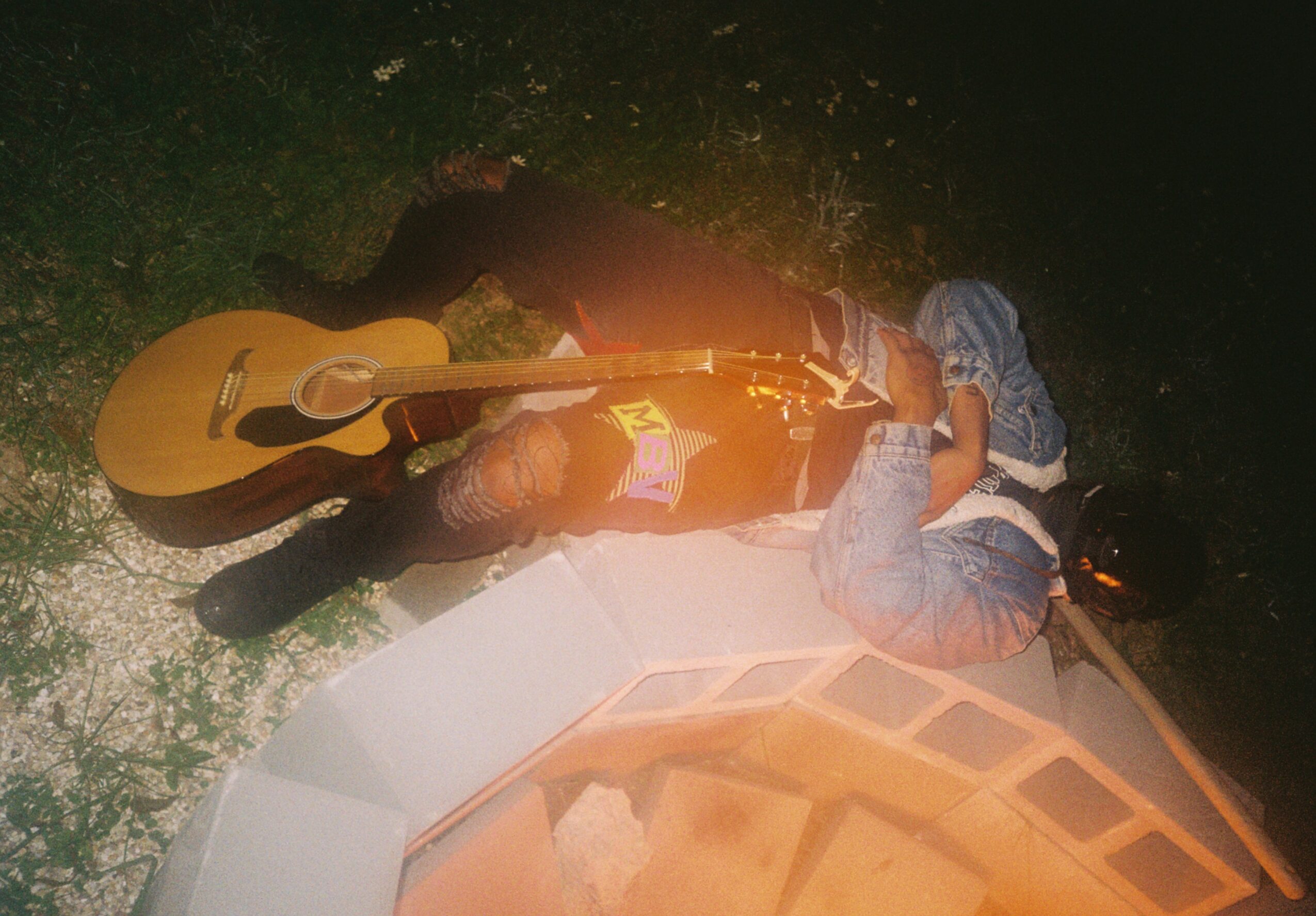 Two people sitting outdoors, one playing guitar, the other leaning on a white structure, at night with dark background.