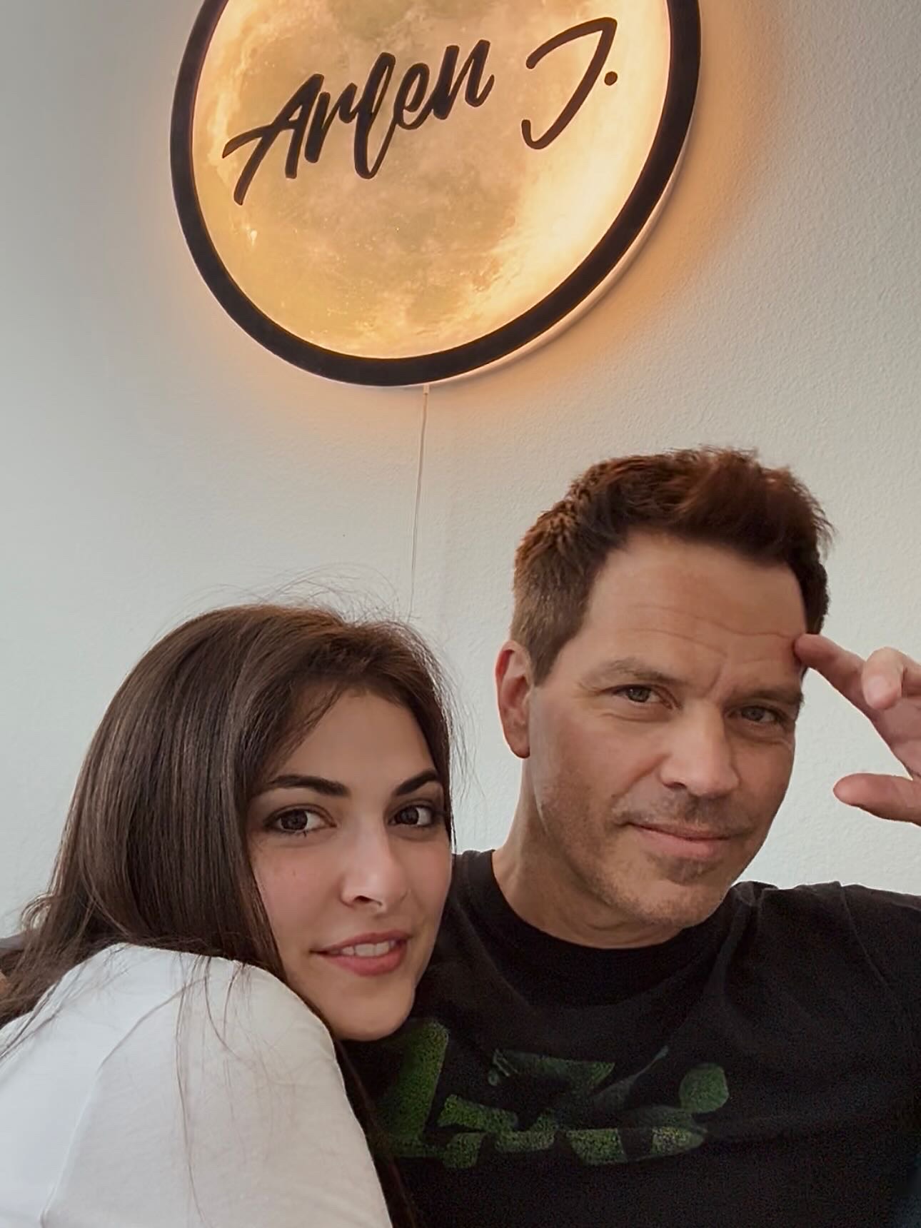 A woman and a man pose together indoors, with a round sign above them reading 'Arlen J.' in cursive.