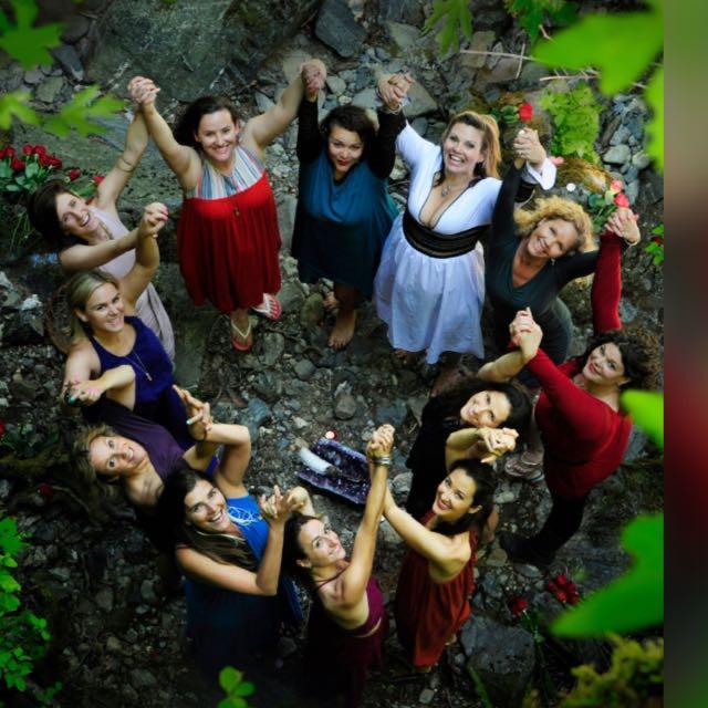 Group of people holding hands in a circle outdoors, smiling, surrounded by rocks and greenery.