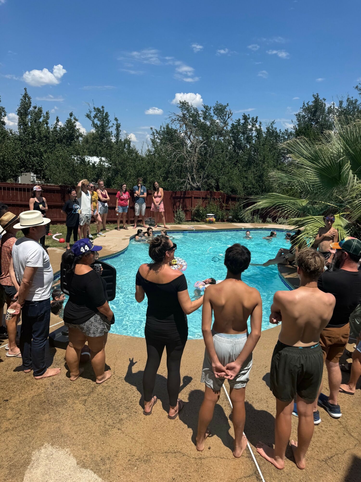 People gathered around a swimming pool on a sunny day, some standing and some sitting, with trees and blue sky in background.