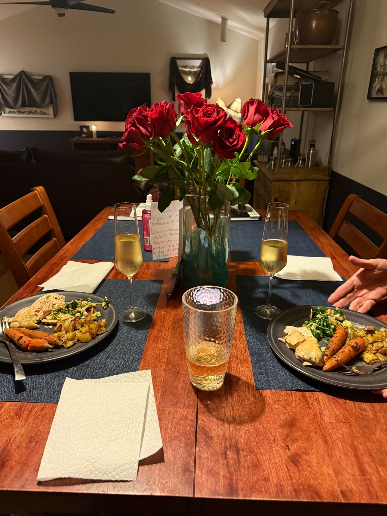 Dining table with a vase of red roses, two plates of food, and drinks, set in a cozy room.