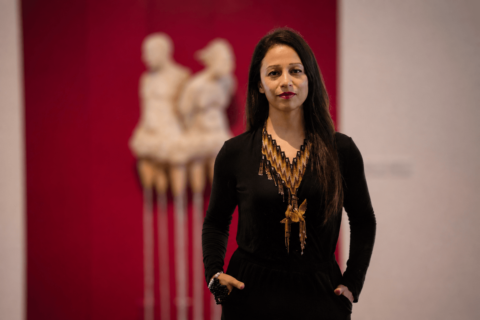 Woman with long dark hair wearing a black outfit and gold jewelry, standing in an art gallery with sculptures in the background.