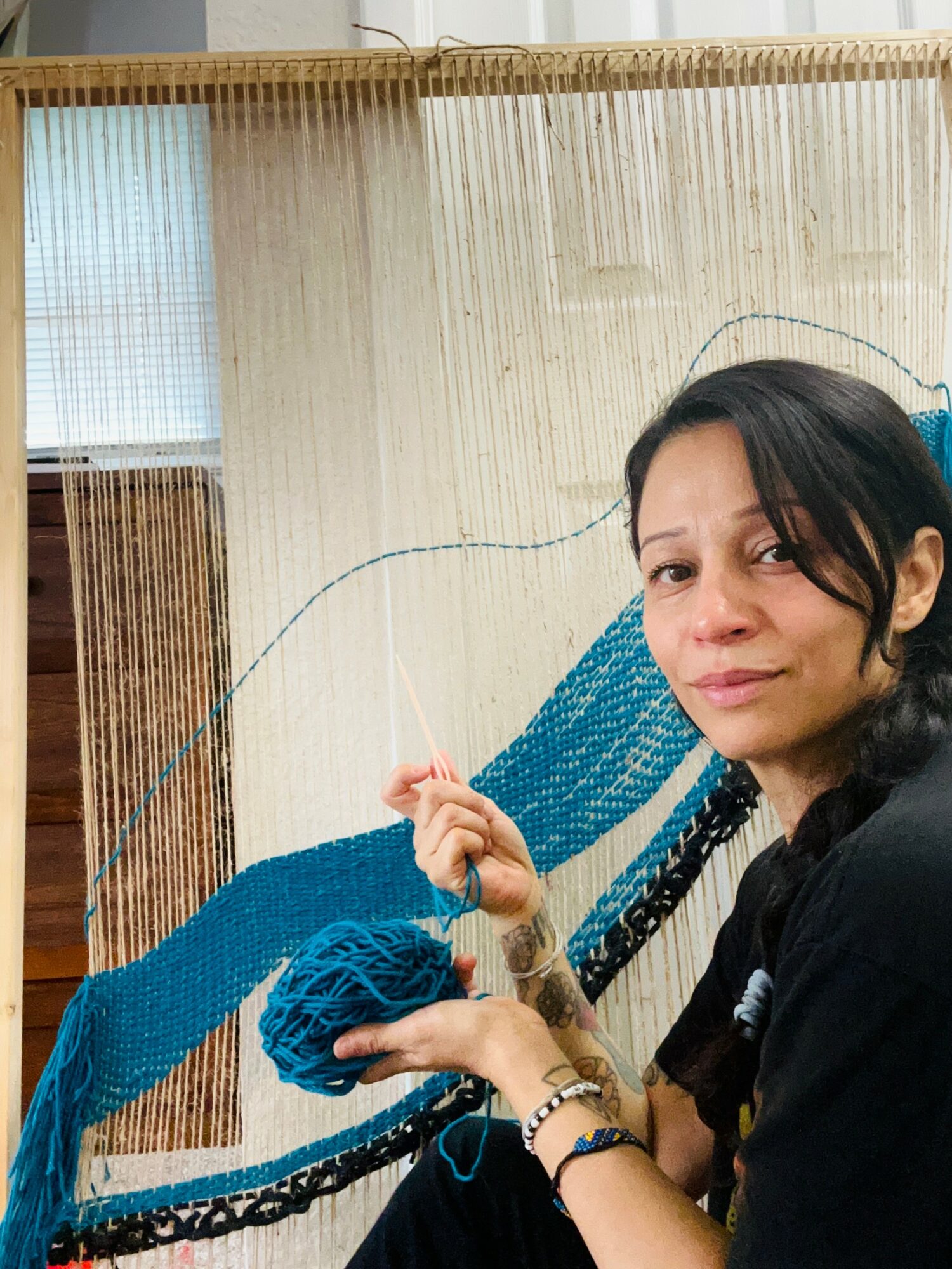 Woman with dark hair holding blue yarn, sitting in front of a woven background, smiling slightly.