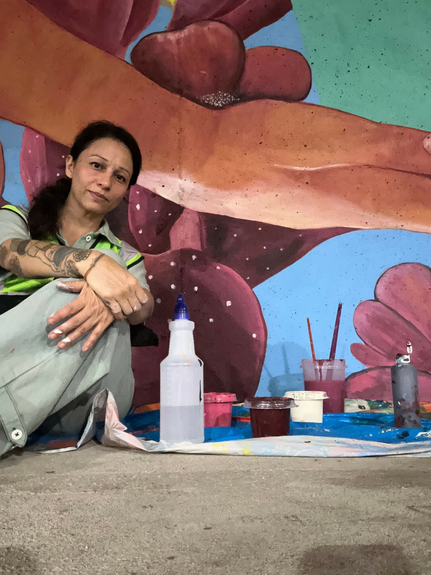 Person with long dark hair and tattoos sitting at a table with art supplies, colorful mural background.