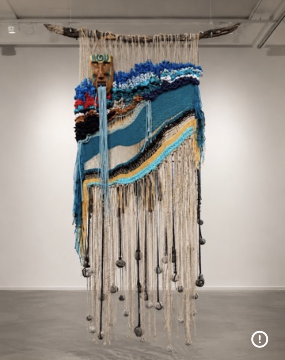 Textured wall hanging with blue, black, and beige yarns, featuring waves and hanging tassels, displayed on a white gallery wall.