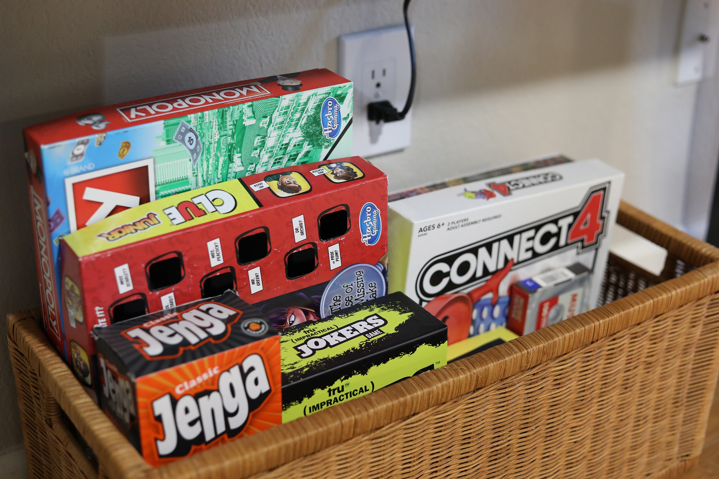 A wicker basket containing board games and puzzles on a table, with a wall and electrical outlets in the background.