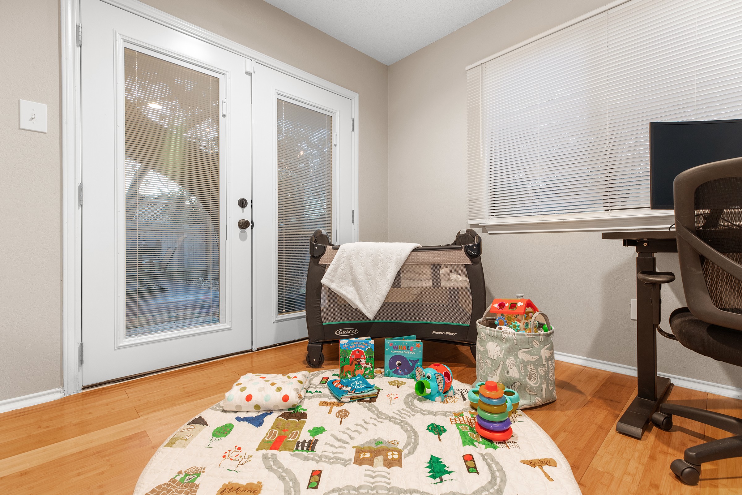 Room with door, window, desk, chair, and colorful toys on a rug, with a blanket on a bench.