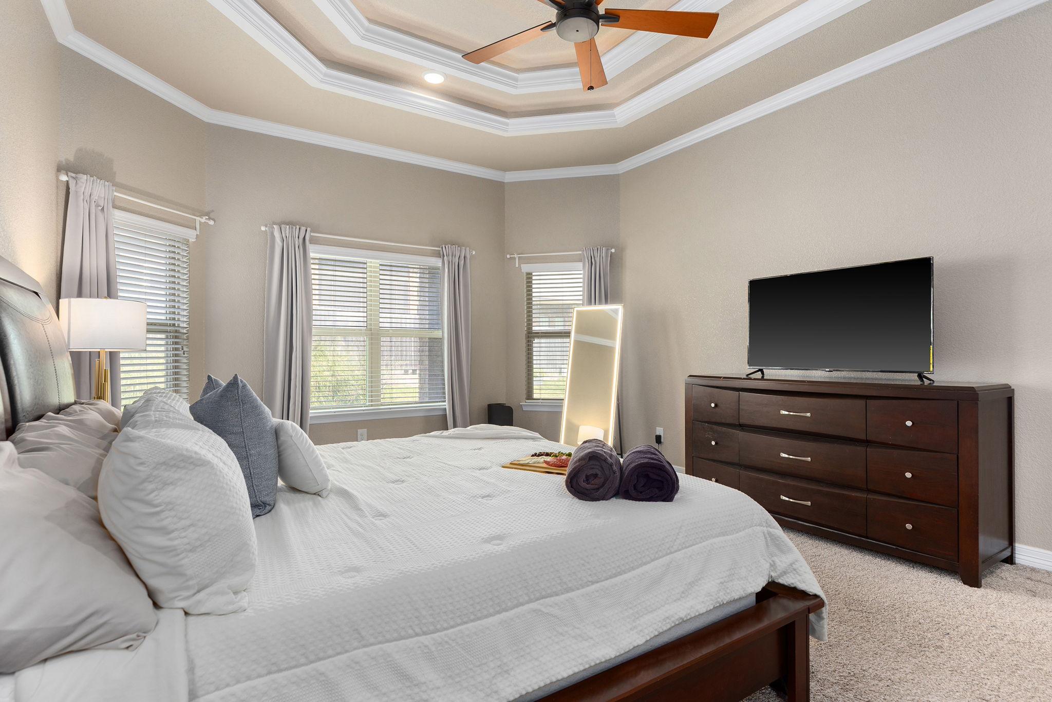 Bedroom with a large bed, multiple pillows, windows with curtains, a dresser with a TV, and a ceiling fan.
