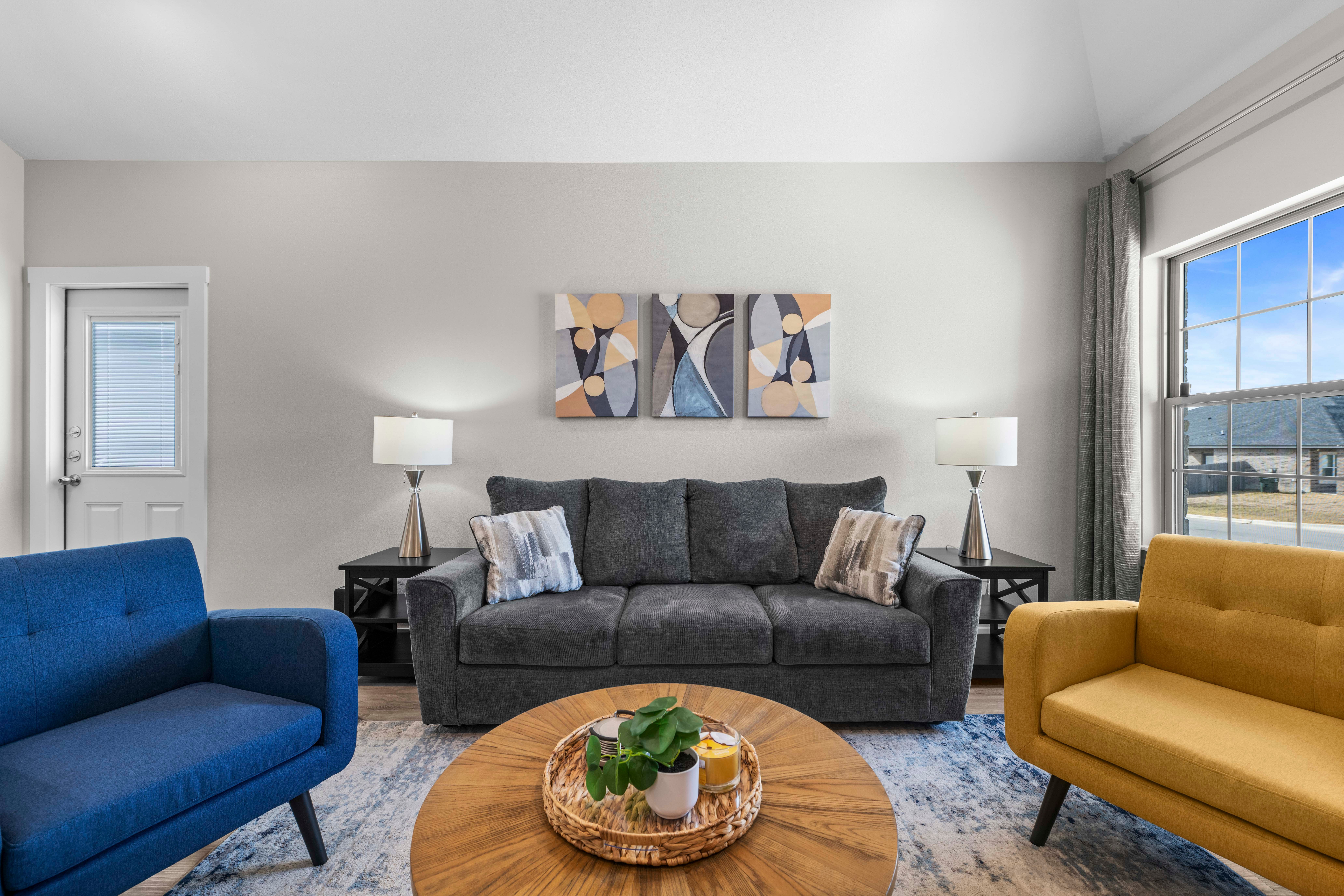 Living room with a gray sofa, two side tables with lamps, a round wooden coffee table, and colorful armchairs, large window, and artwork.