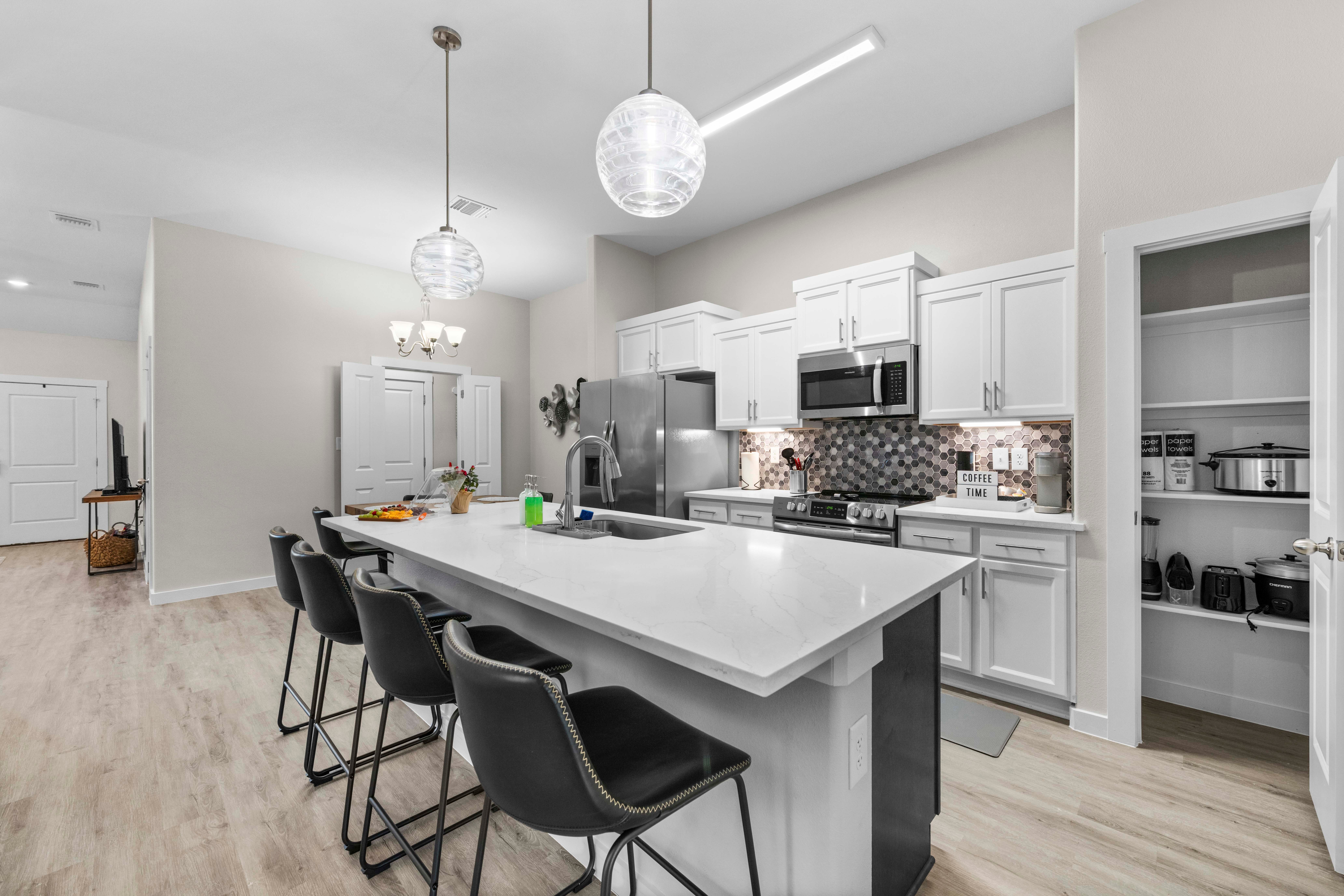 Modern kitchen with white cabinets, island, black chairs, stainless steel appliances, and pendant lighting, open to dining and living areas.