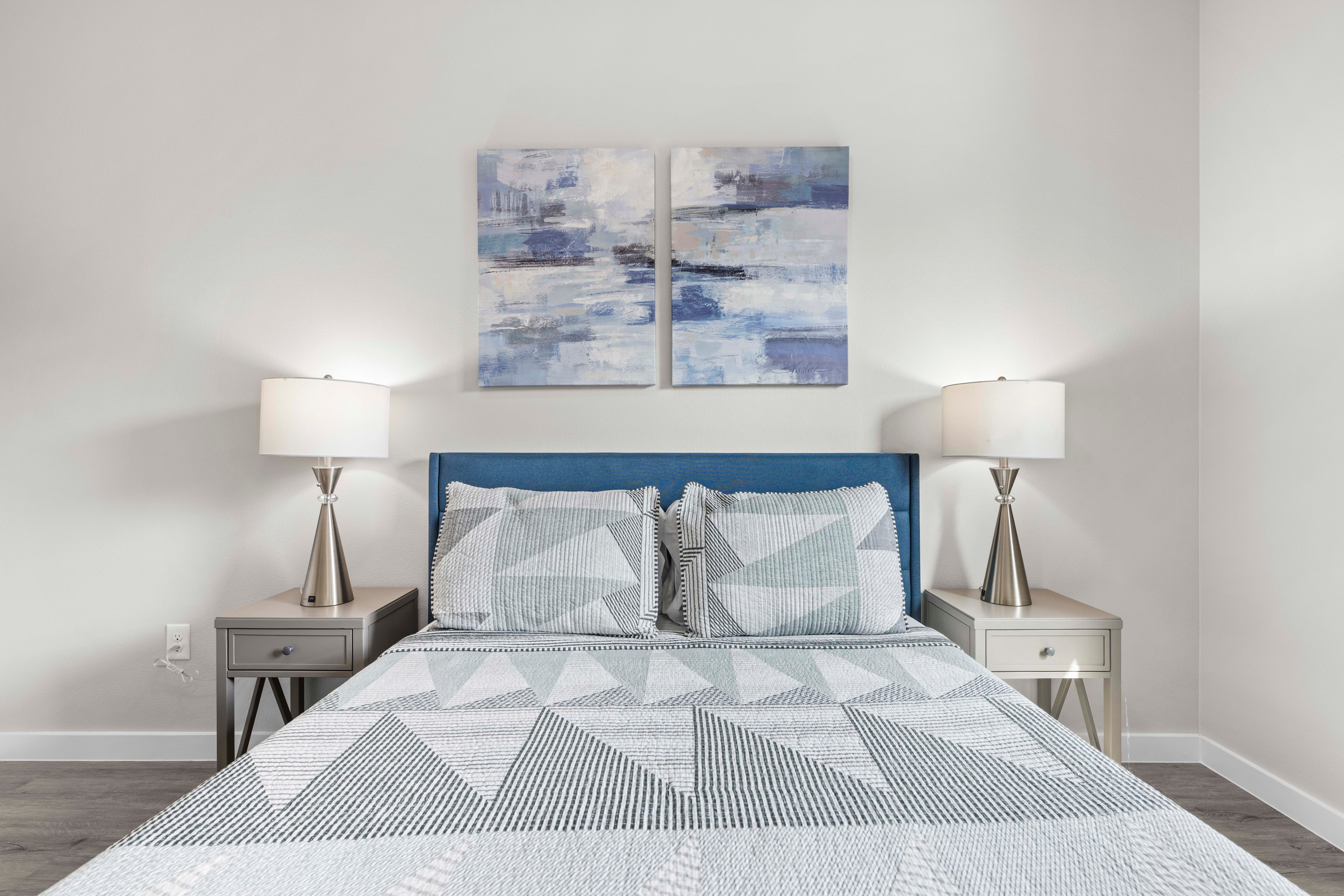 Upright view of a bedroom with a bed, two nightstands, lamps, and abstract artwork above the headboard.
