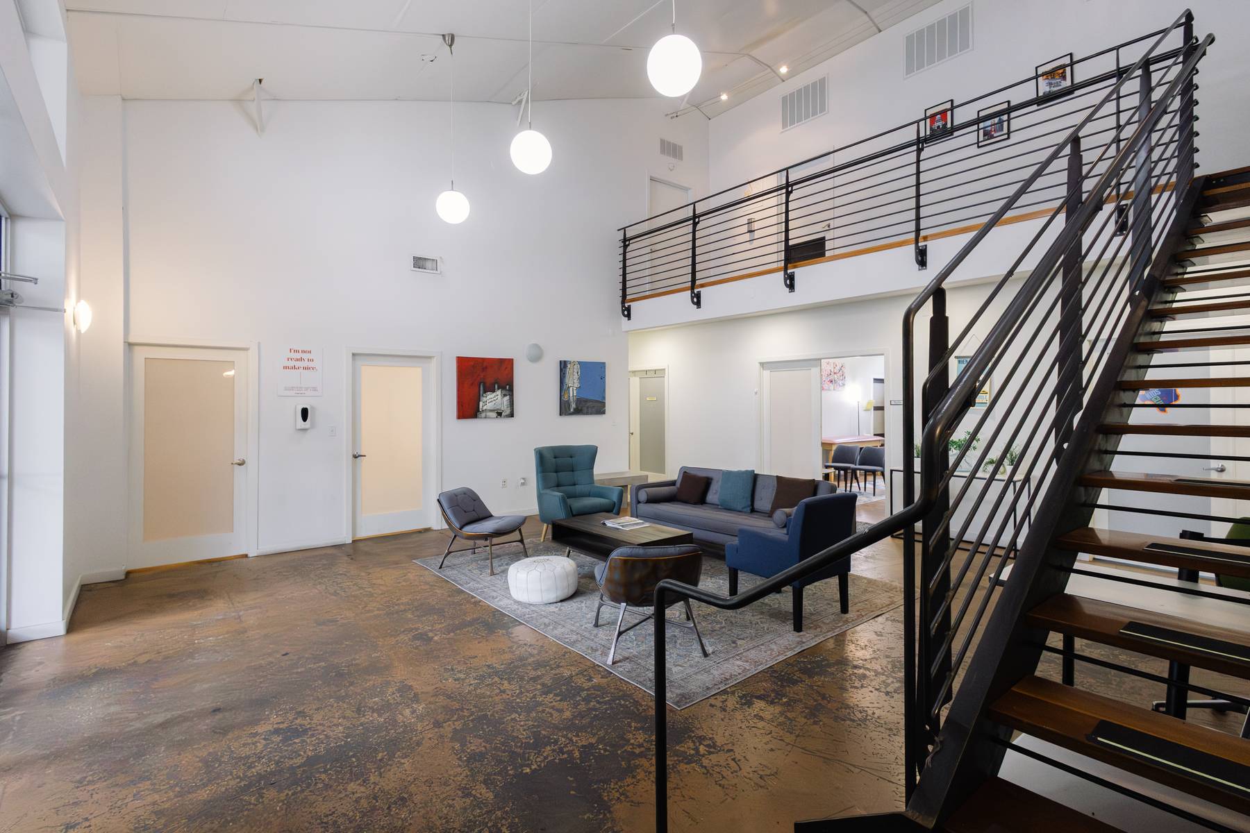Modern interior with high ceiling, seating area with sofa and chairs, artwork on white walls, staircase with black railing, and hanging lights.