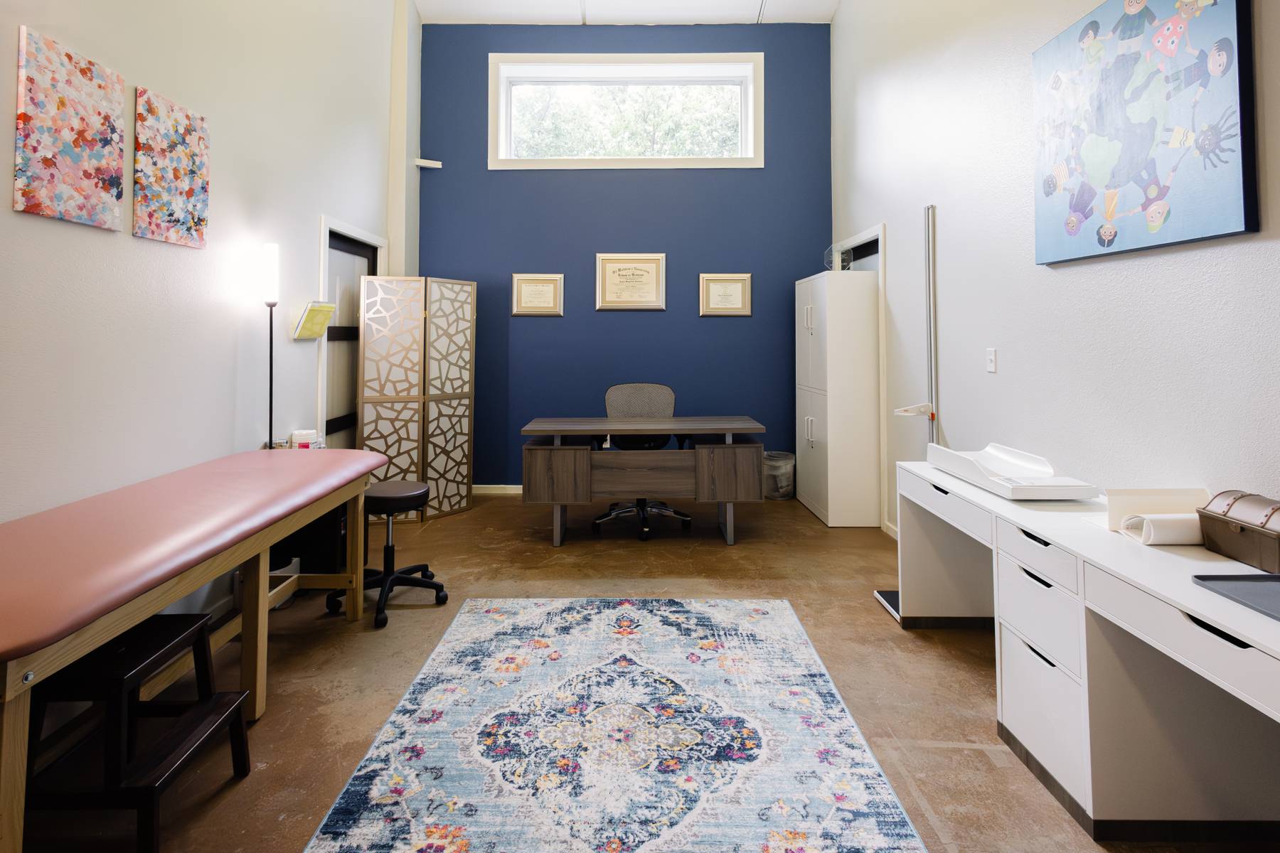 Medical examination room with a pink examination table, white cabinets, a rug, and framed artwork on the walls.