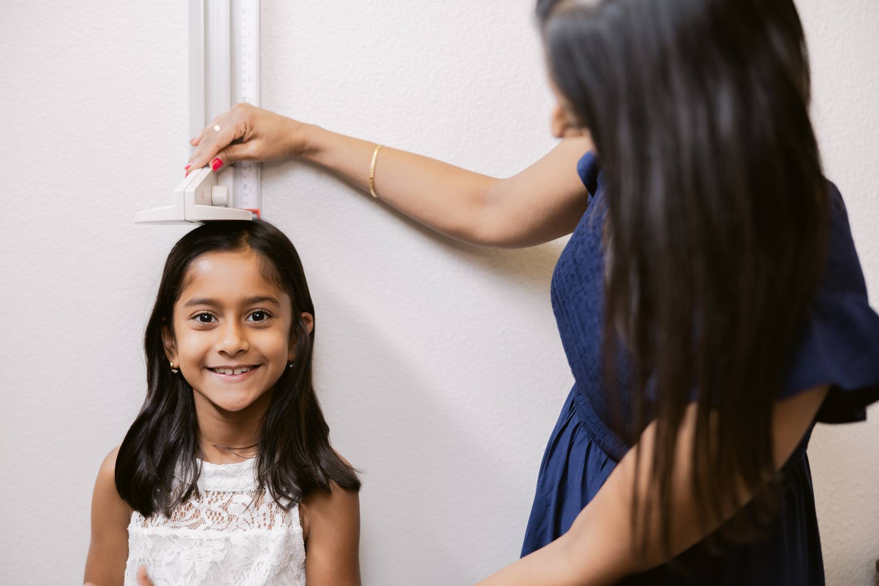 Young girl with long dark hair smiling, standing near a wall, with an adult woman adjusting a light switch above her.