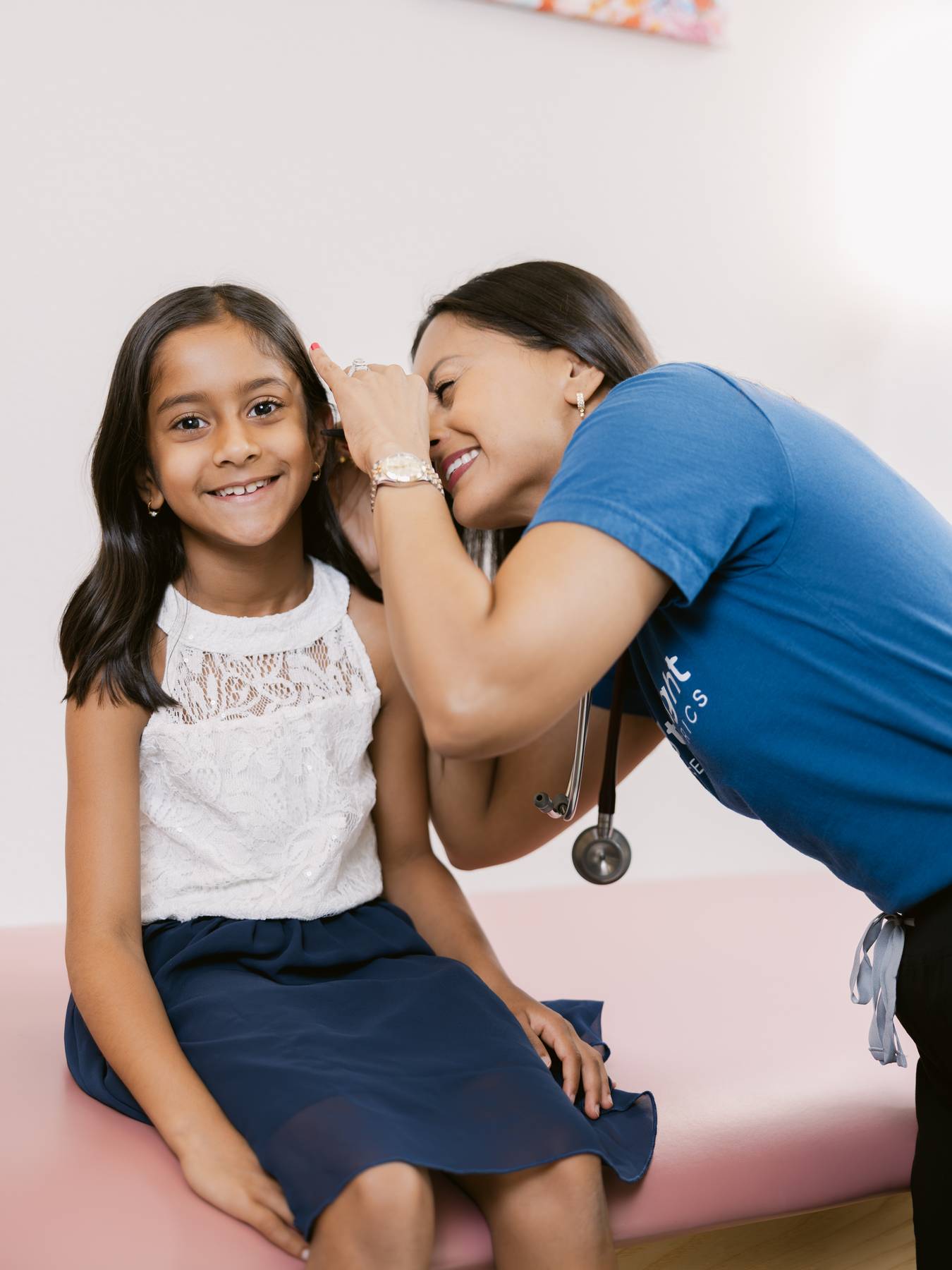 A healthcare worker whispers to a smiling young girl sitting on an examination table, both smiling and engaging happily.