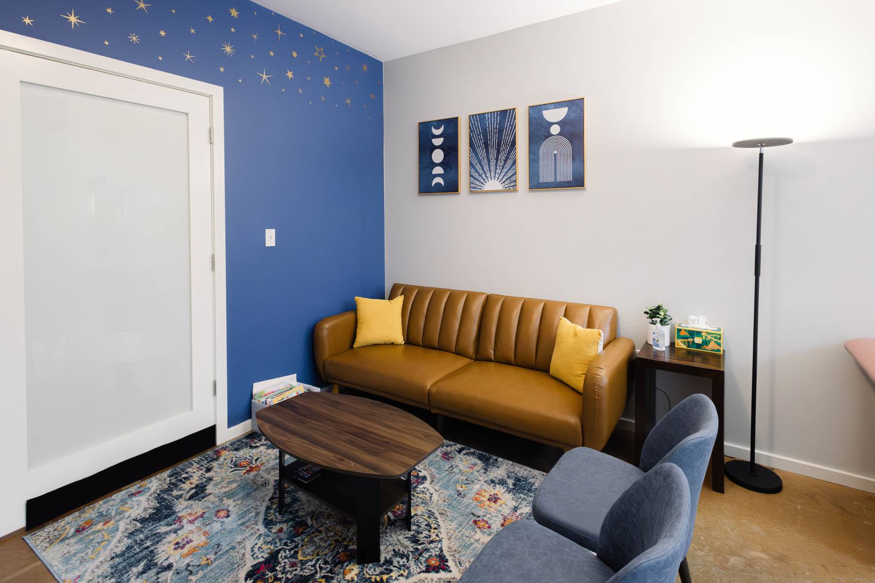 Living room with a blue accent wall, a mustard yellow sofa, three wall art pieces, a floor lamp, and a patterned rug.