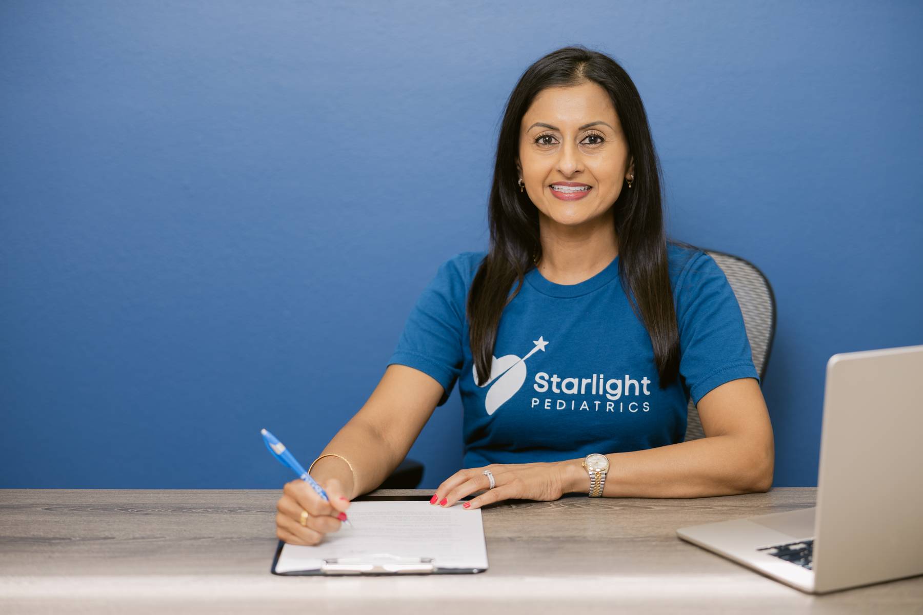Woman sitting at desk with notebook, smiling, wearing a blue Starlight Pediatrics shirt, against a blue background.