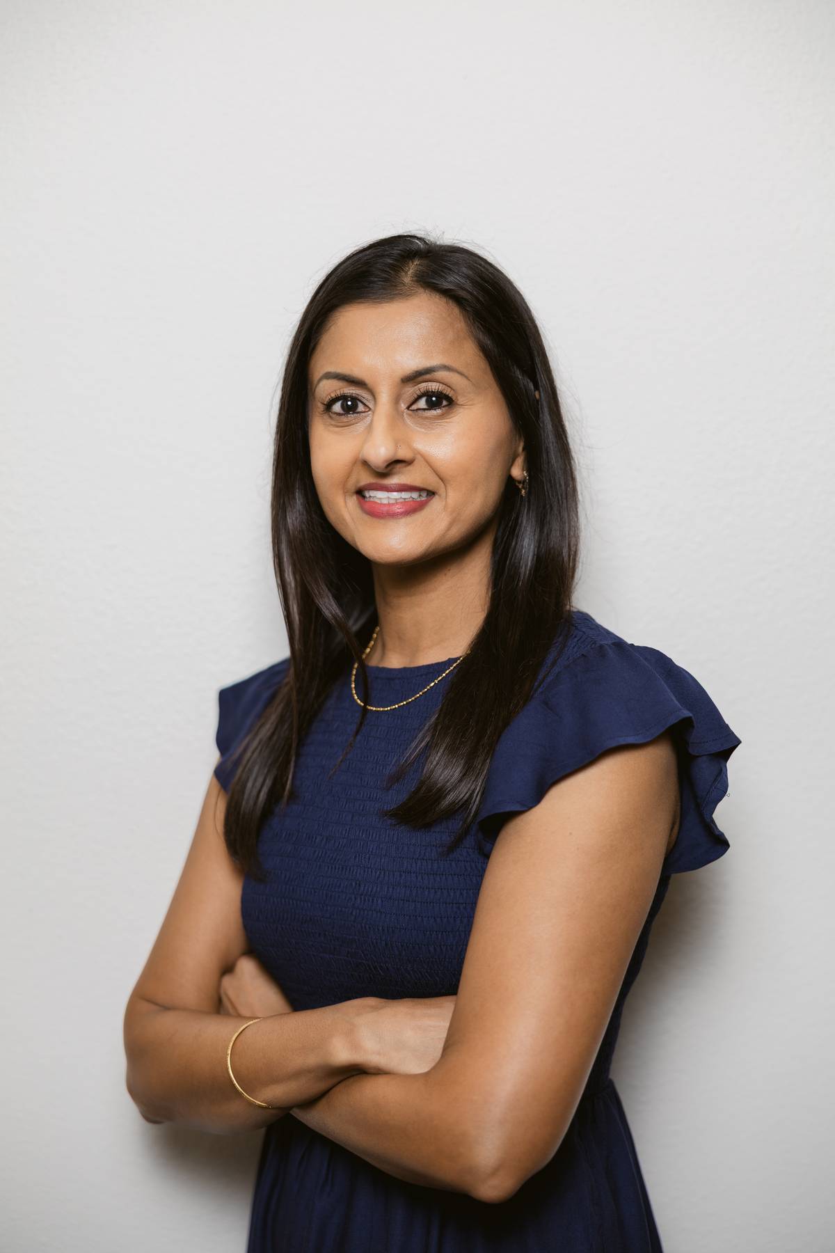 Woman with long dark hair wearing a navy blue dress, smiling with arms crossed against a plain white background.