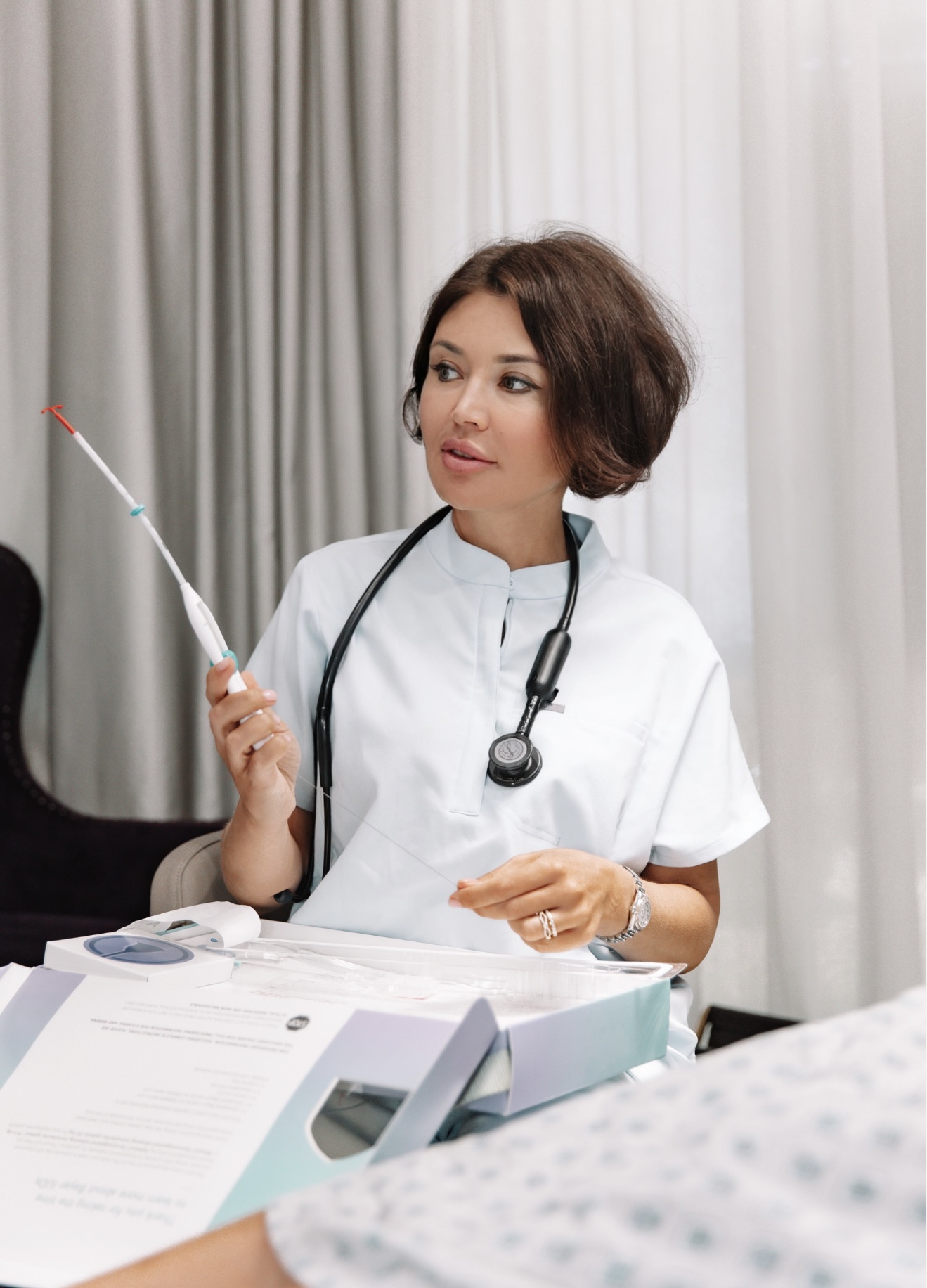 Female healthcare professional with a stethoscope around her neck, holding a syringe, sitting at a desk with papers and medical equipment.