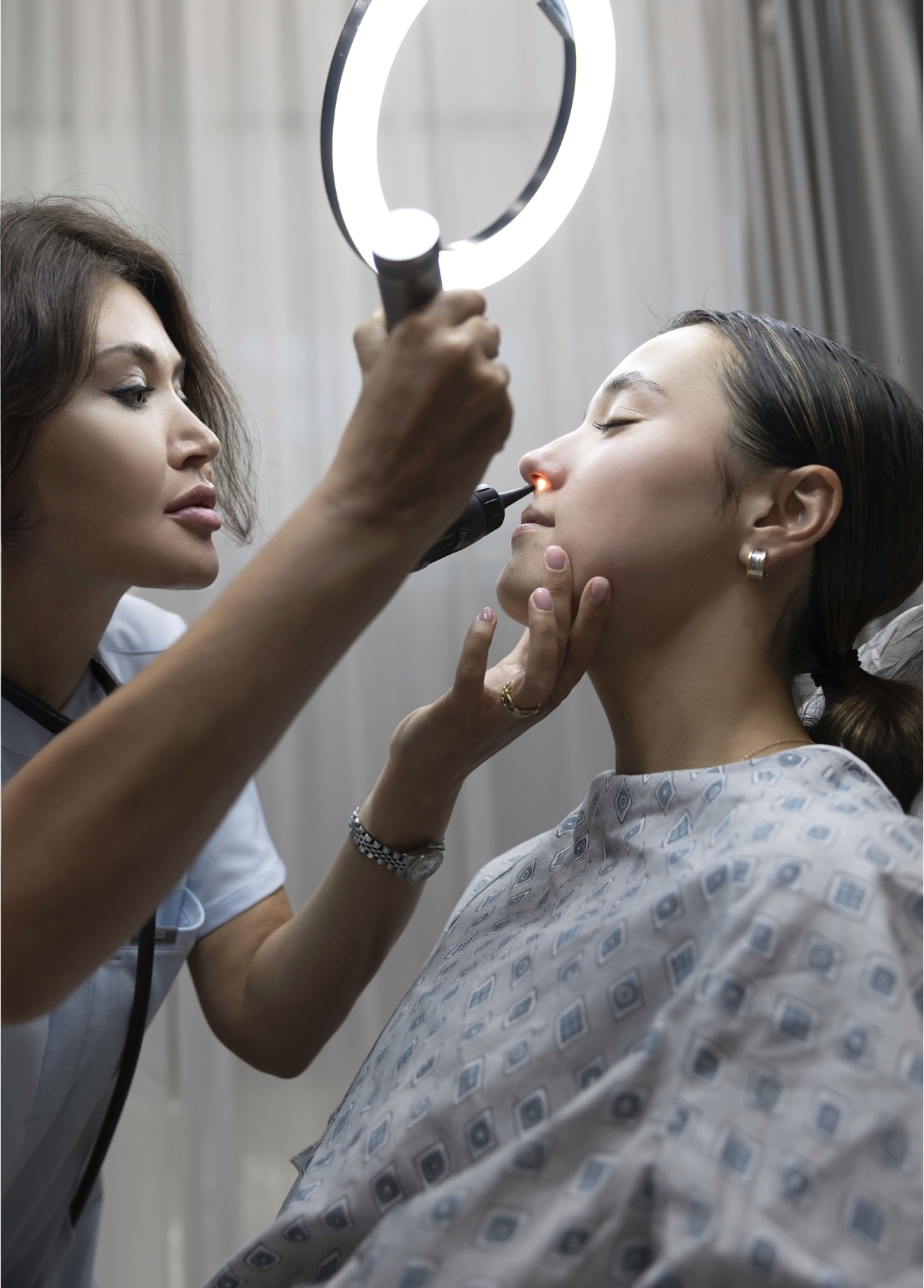 Makeup artist applies makeup to a woman's face in a clinical setting with a ring light, woman in hospital gown.