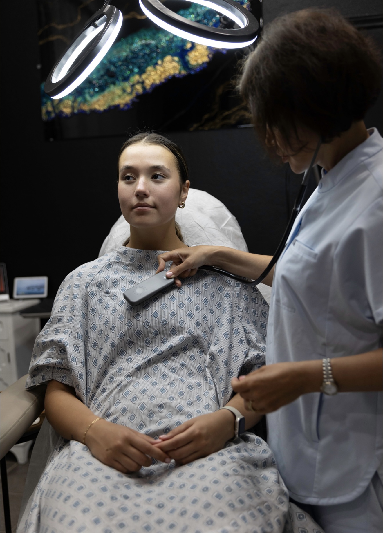 Young woman in hospital gown sitting on examination table with healthcare professional beside her, in a medical setting.
