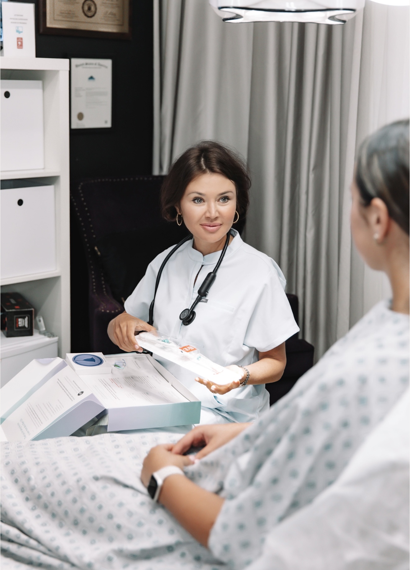 Female doctor talking to a patient in a hospital room, holding a clipboard, with medical certificates on the wall.