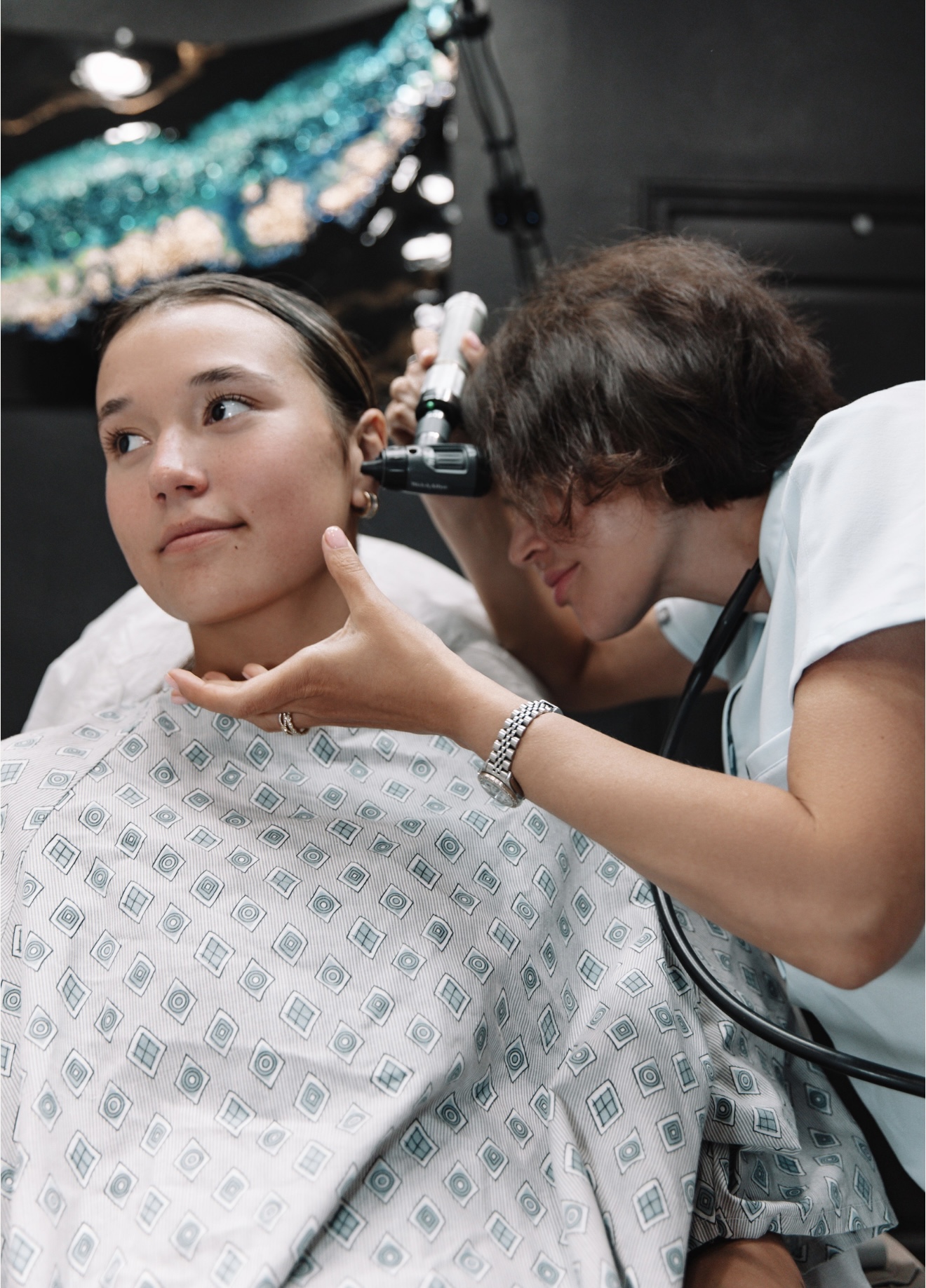 A woman examines a young woman's face in a medical setting, with decorations and equipment visible in the background.