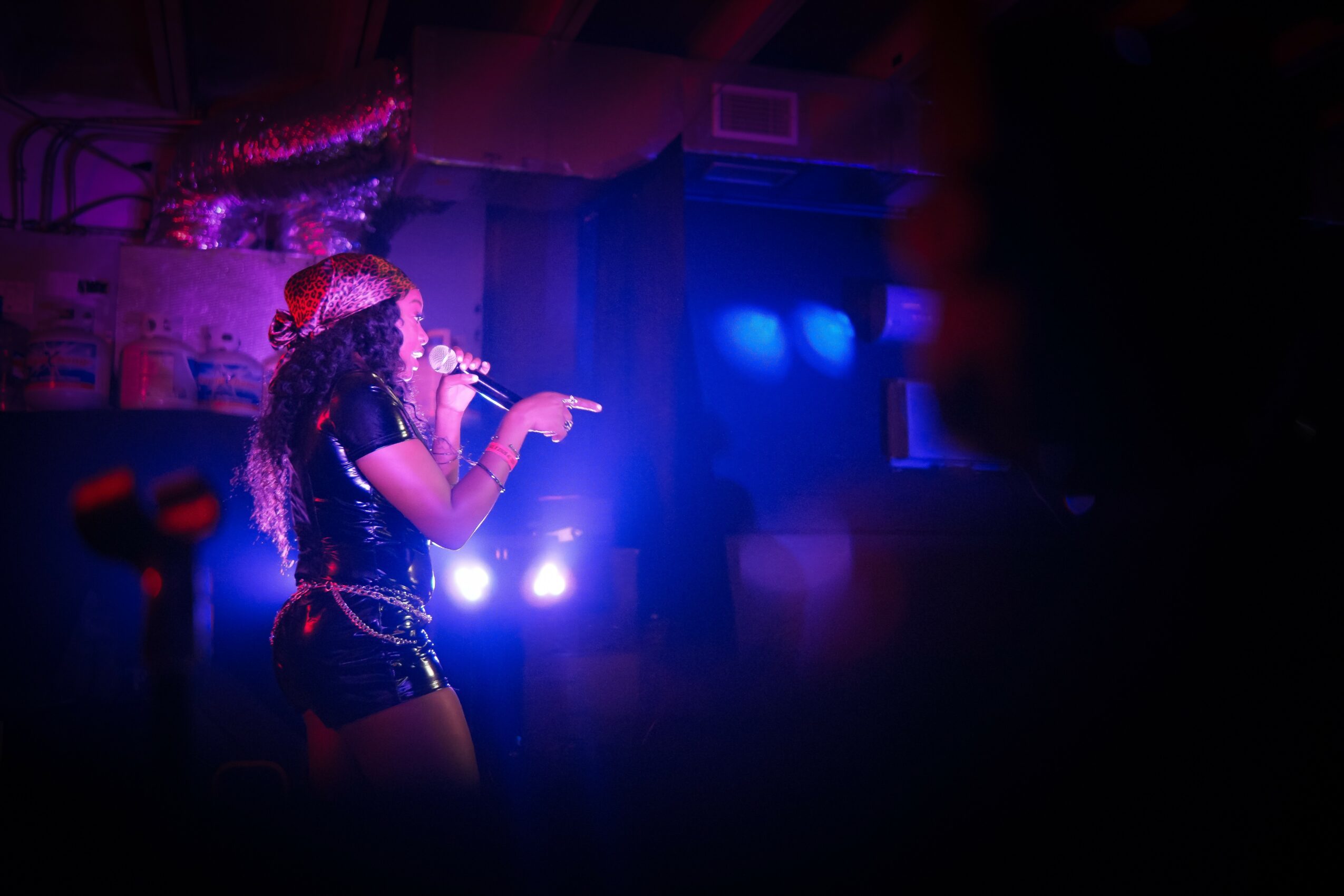 Person singing or speaking into a microphone on stage with colorful lighting, facing left, in a dark setting.