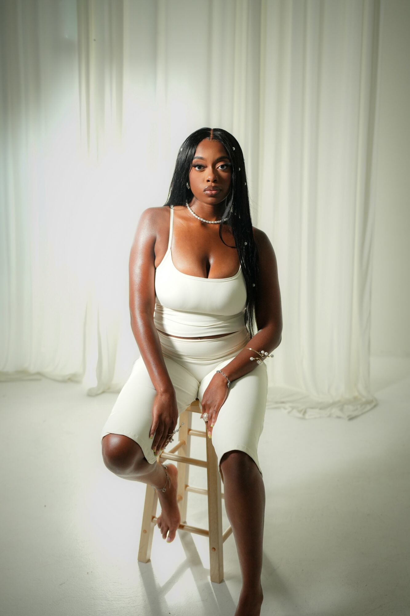 Woman with long black hair sitting on a wooden stool in front of light-colored curtains, wearing a white dress.