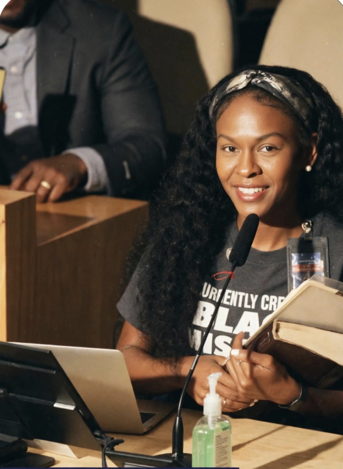 Young woman with curly hair smiling, holding a book, sitting at a desk with a microphone and sanitizer, in a meeting.