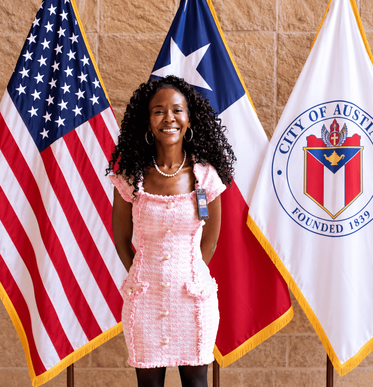 Smiling woman standing in front of American, Texas, and Austin city flags, wearing a pink dress and pearl necklace.