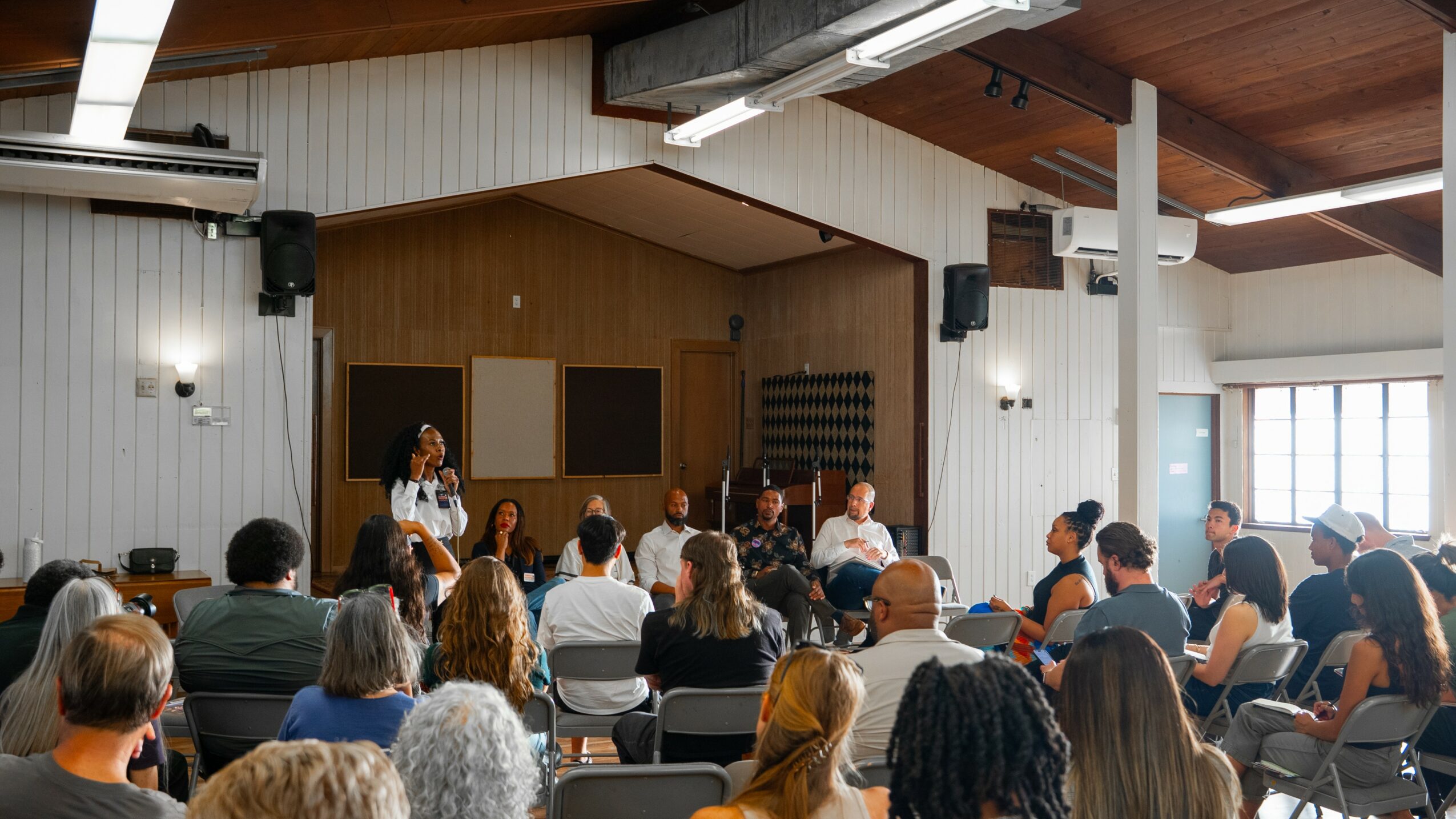 People seated in a room listening to a woman speaking on stage, with a wooden wall and large windows.