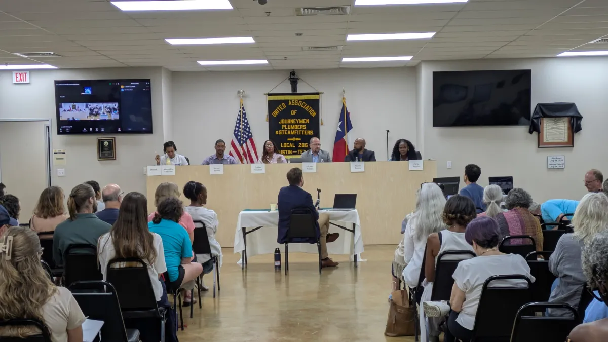 Panel of six people seated at a long table in a room with an audience, American flags, and a black and gold emblem on the wall.