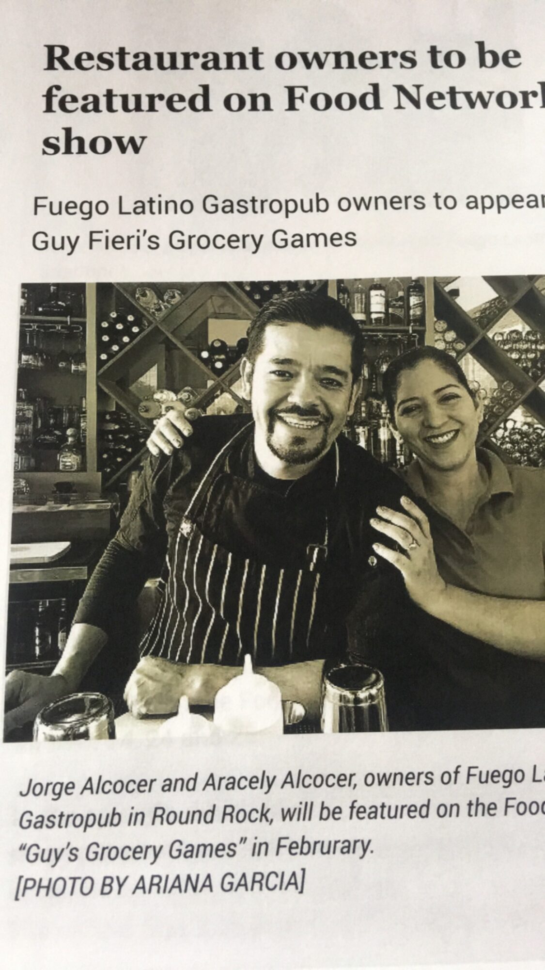 Two people smiling, one wearing an apron, in a restaurant setting with shelves and bottles behind them.