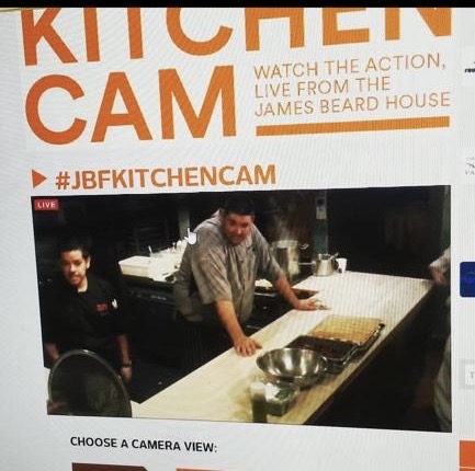 Two people in a kitchen, one leaning on a counter, with cooking utensils and a baking sheet. Text about a kitchen camera event.
