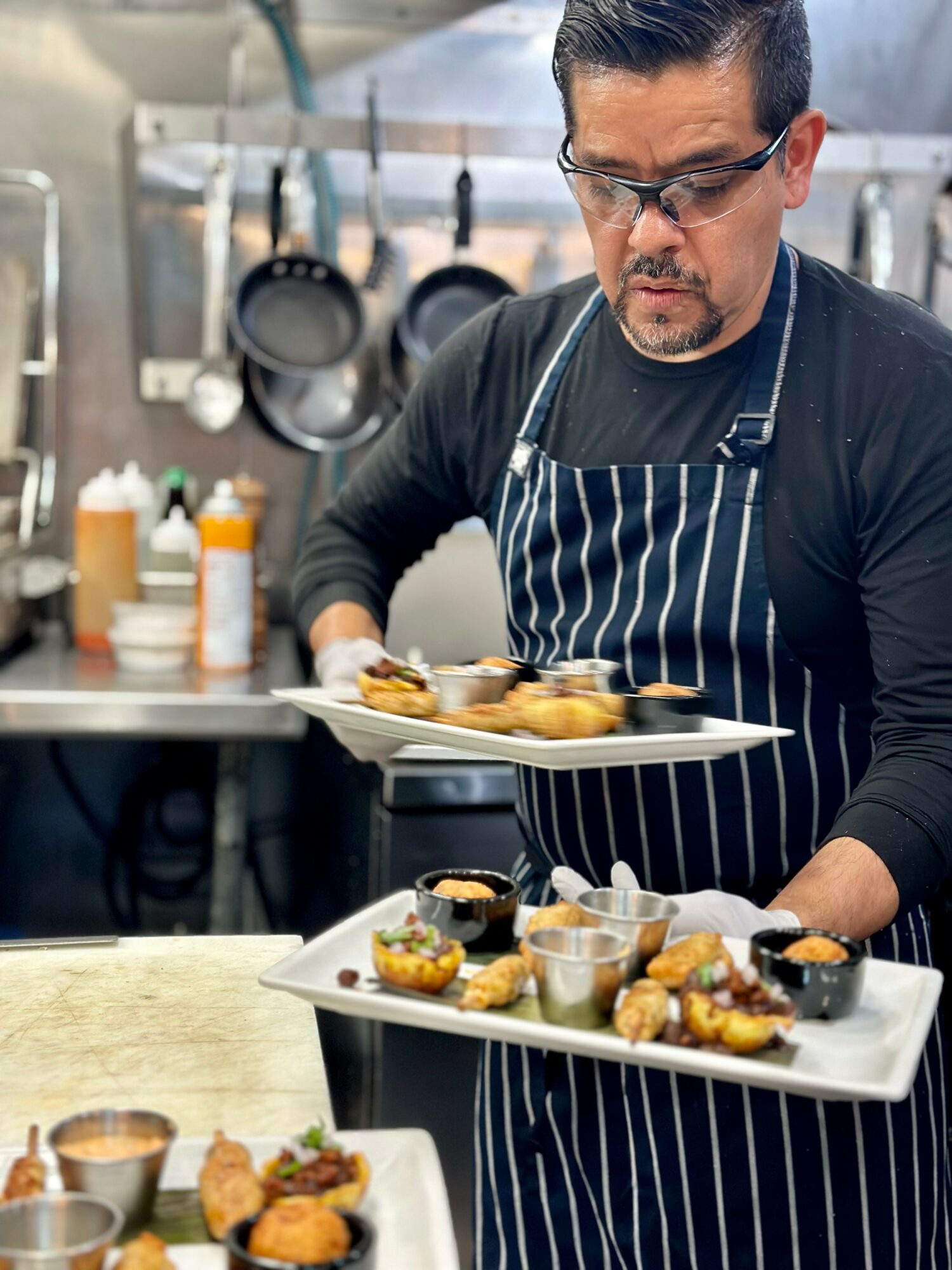 Man in black shirt and striped apron preparing food in a kitchen with utensils and ingredients, holding a tray of appetizers.