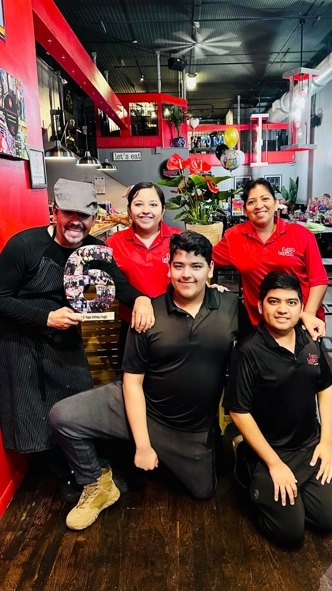 Group of five people smiling inside a restaurant, some wearing red shirts, with plants and decor in the background.