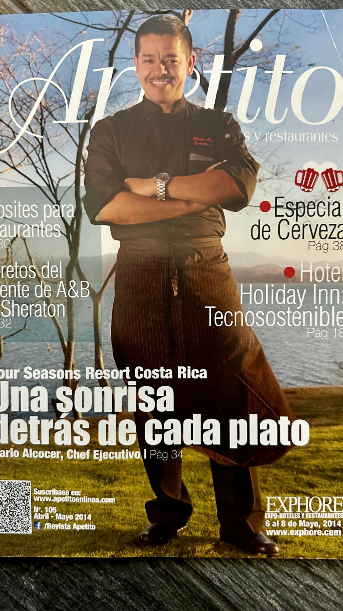 Man in black shirt and brown pants standing outdoors with trees and sky in background, smiling with arms crossed.