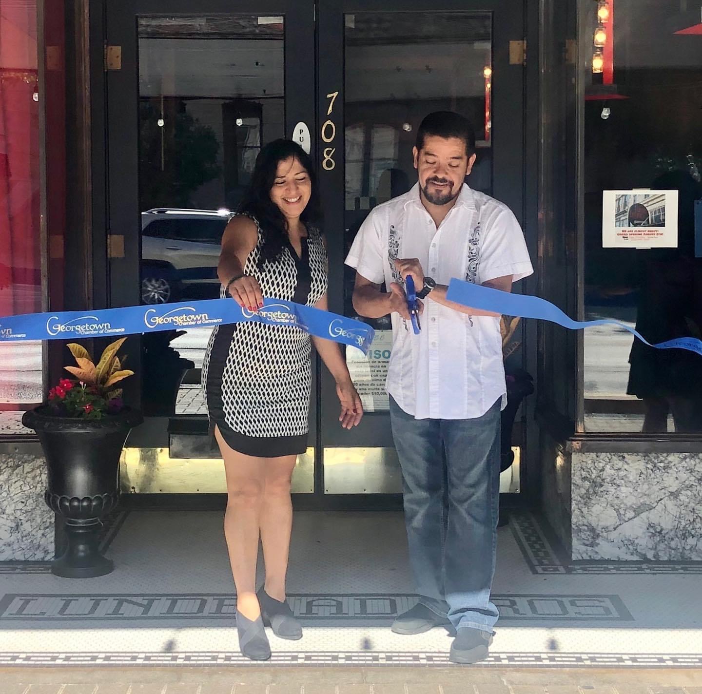 Man and woman at ribbon-cutting ceremony outside building, woman smiling, man holding scissors, blue ribbon, potted plant nearby.