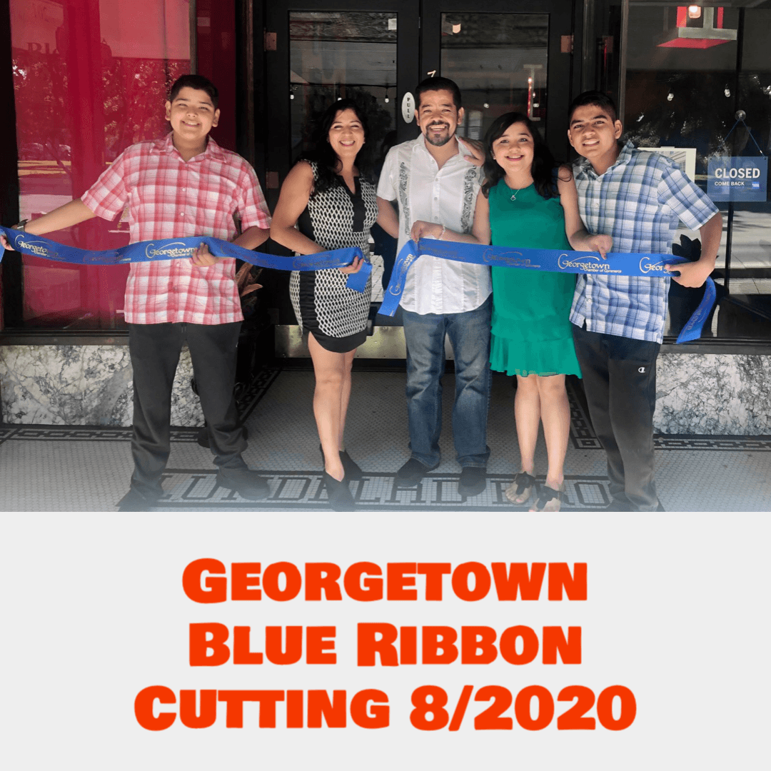 Group of six people standing in front of a store holding a blue ribbon for a ribbon-cutting event, smiling.