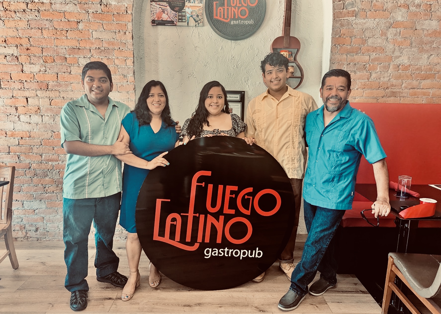 Five people standing indoors behind a round sign that reads 'Fuego Lindo gastropub,' with brick walls and decor in the background.