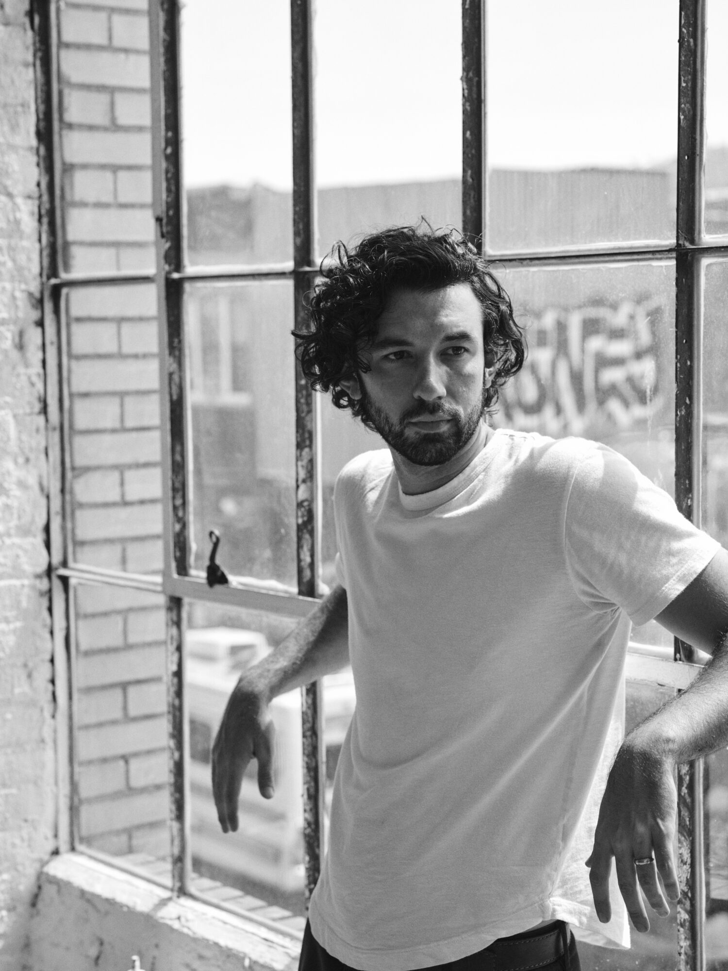 Man with curly hair and beard leaning on window bars, wearing a white t-shirt, in black and white photo.