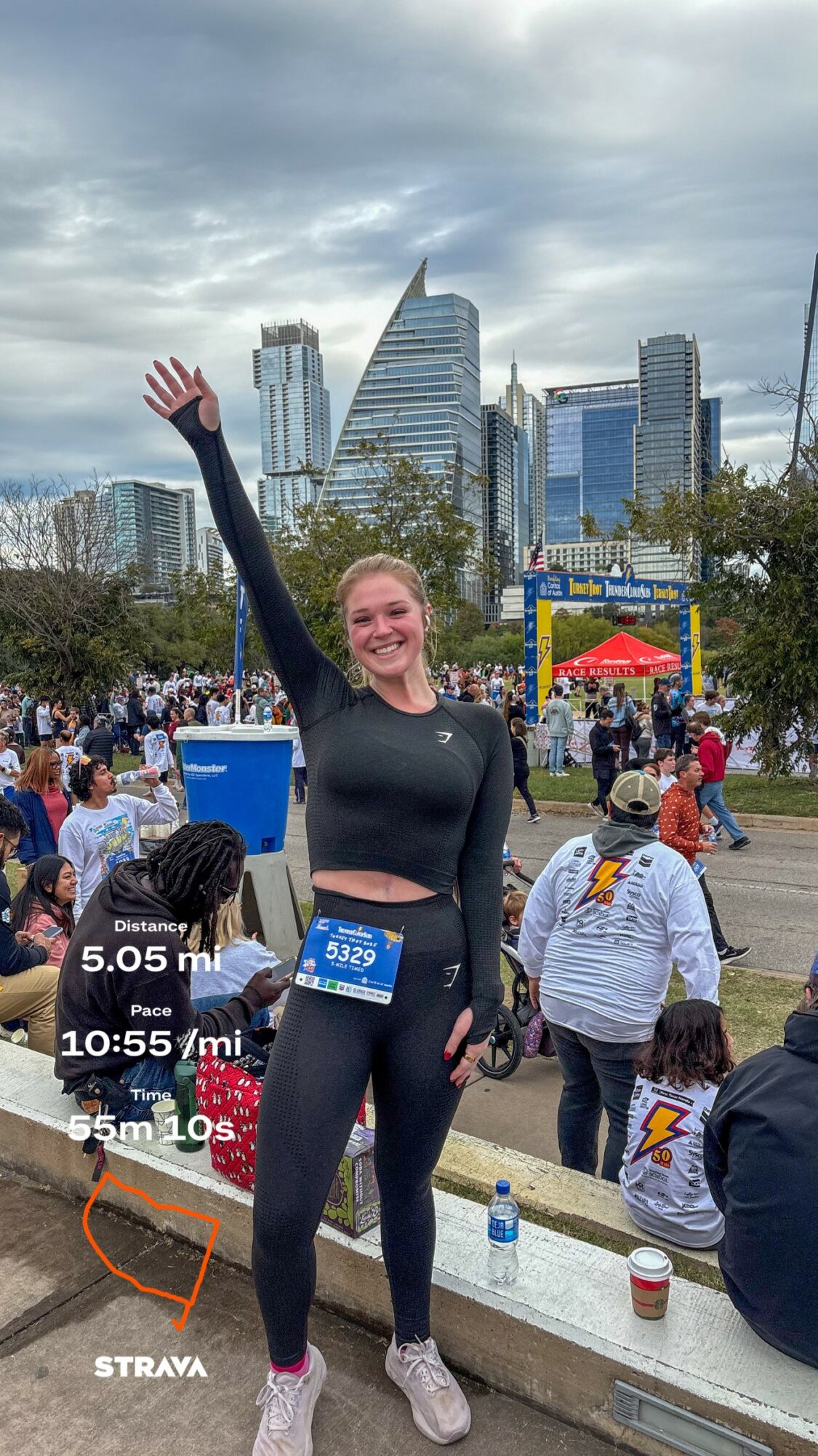 Smiling woman in black athletic clothing raising her arm at outdoor event with city skyline in background.
