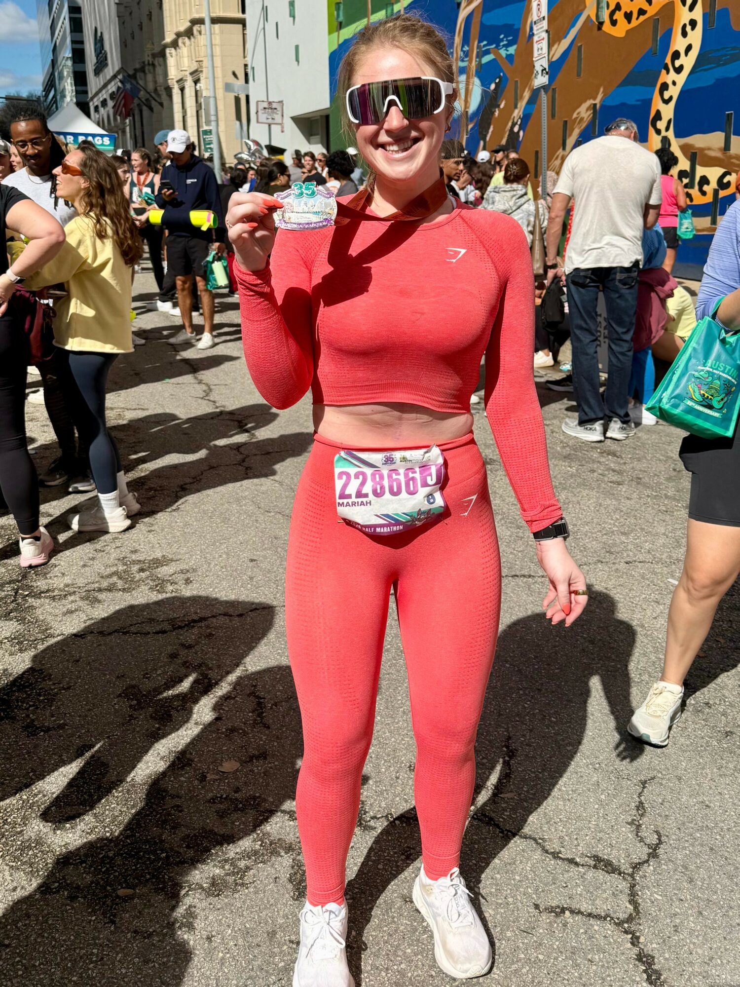 Smiling woman in red athletic outfit holding a medal at outdoor event with crowd and colorful structures in background.