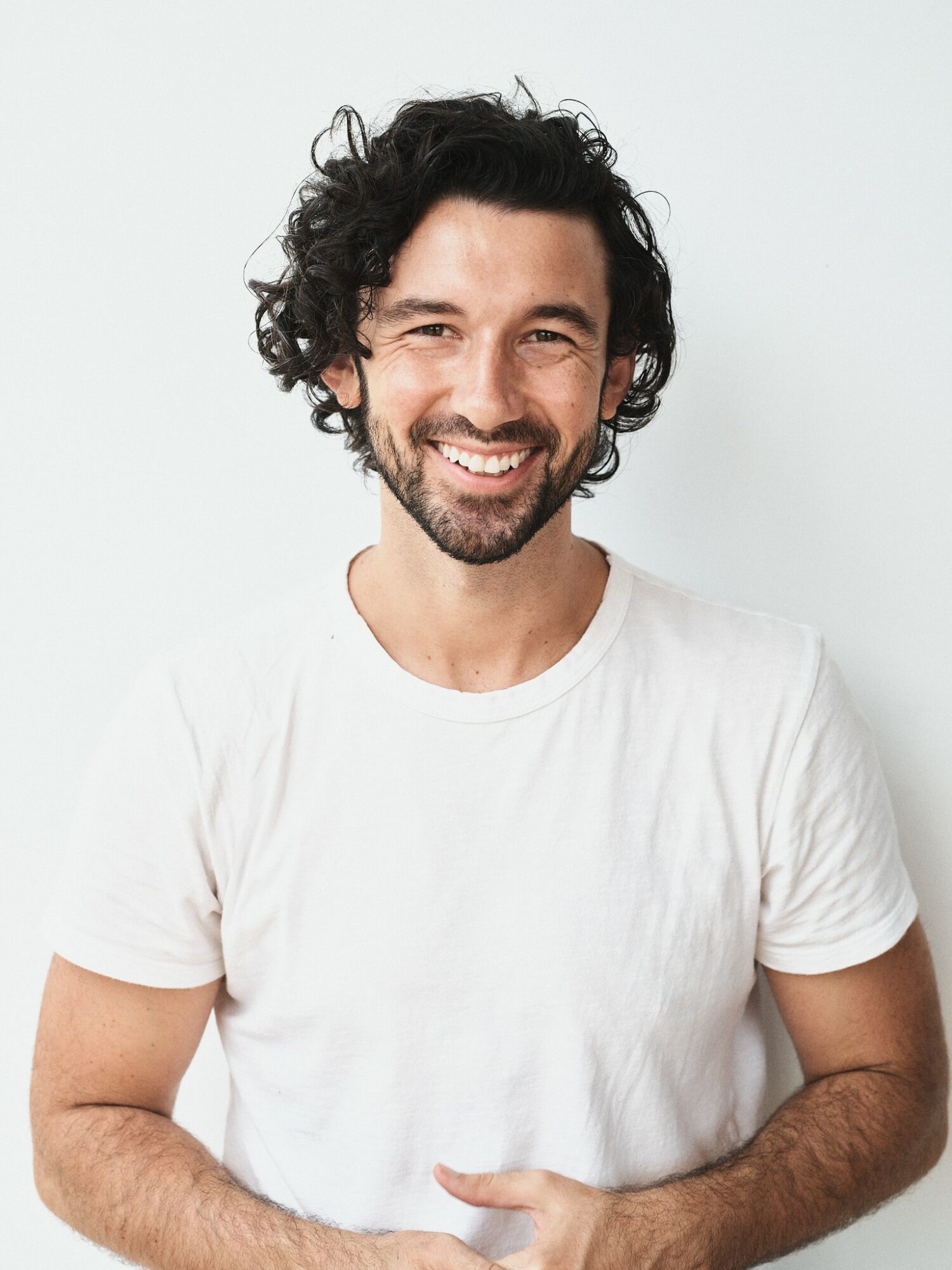 Smiling man with curly dark hair and beard wearing a white t-shirt, standing against a plain light background.