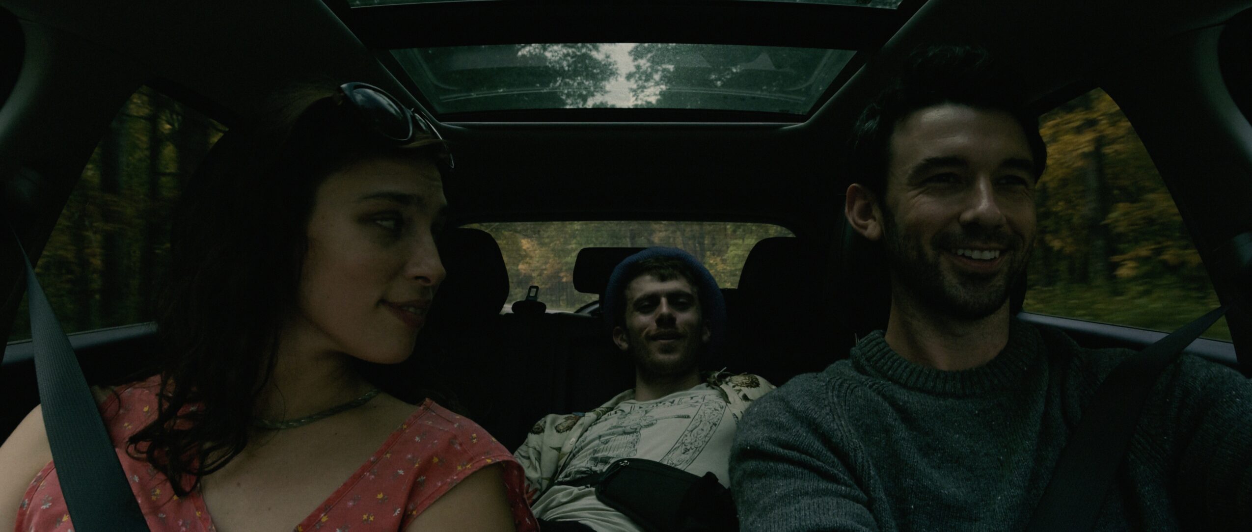 Four people sitting in a car, with trees visible outside the windows, smiling and looking forward.