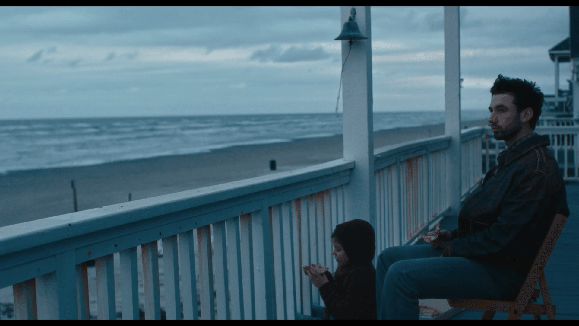 A man and a child sit on a porch overlooking the ocean, with cloudy sky and waves in the background.