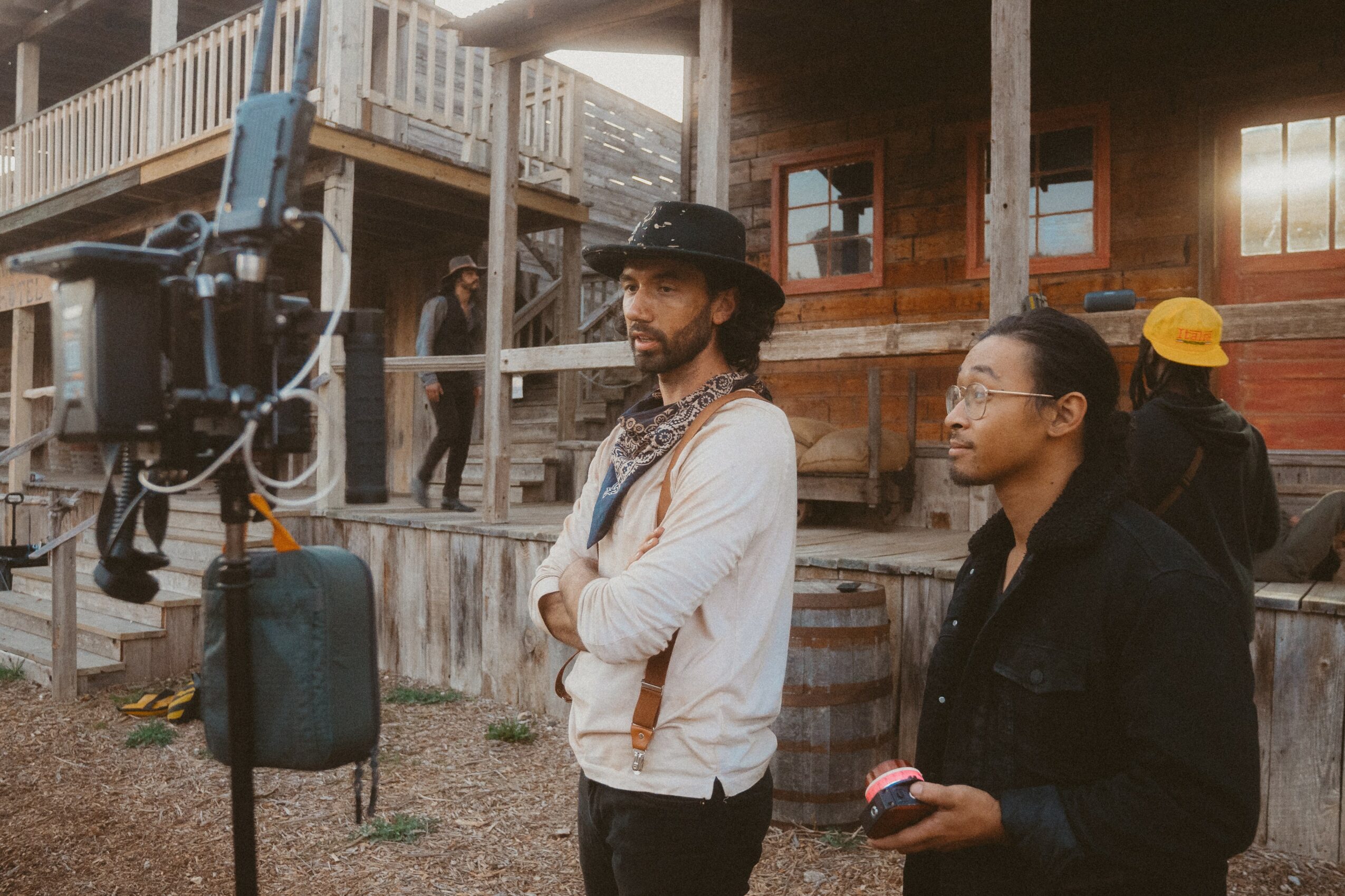 Two people standing outdoors near a wooden building, one wearing a hat and the other holding a phone, with a camera on a tripod nearby.