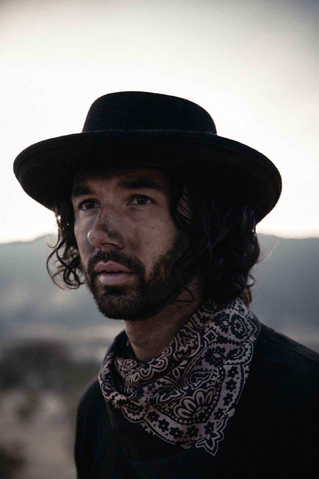 Man with long hair, beard, wearing a wide-brimmed hat and patterned bandana, outdoors with a cloudy sky.